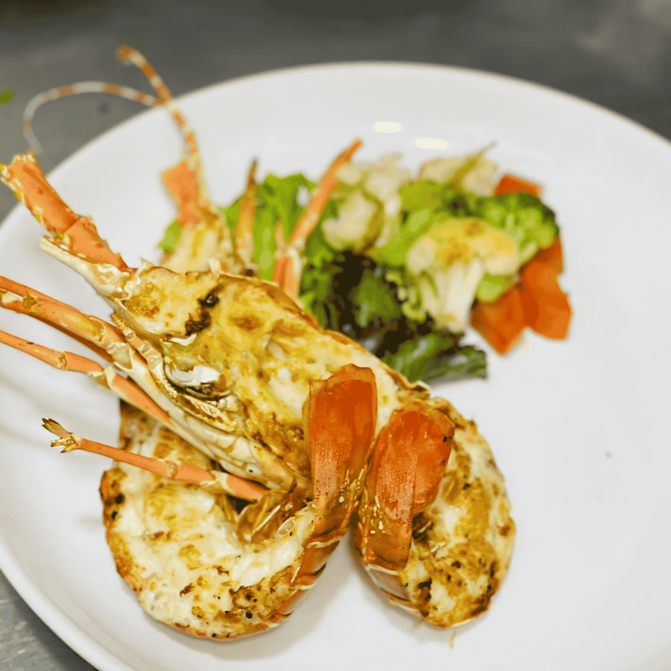 Grilled lobster halves with charred meat, served on a white plate with a small side salad on a metal countertop.